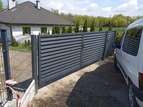 Nowoczesna, pozioma brama przesuwna w kolorze grafitowym, z widocznym fragmentem ogrodzenia panelowego i białym samochodem dostawczym w tle domu jednorodzinnego.