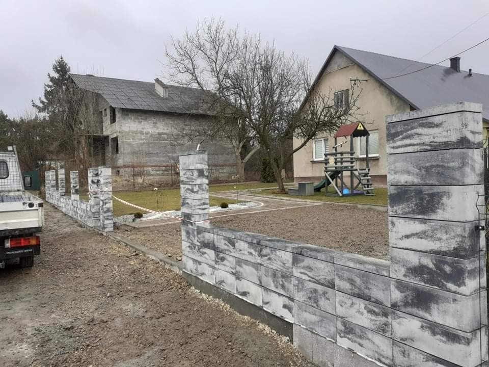 Nowoczesne ogrodzenie z betonowych bloczków imitujących kamień, widoczne na tle domu w budowie i placu zabaw, z samochodem dostawczym zaparkowanym obok.