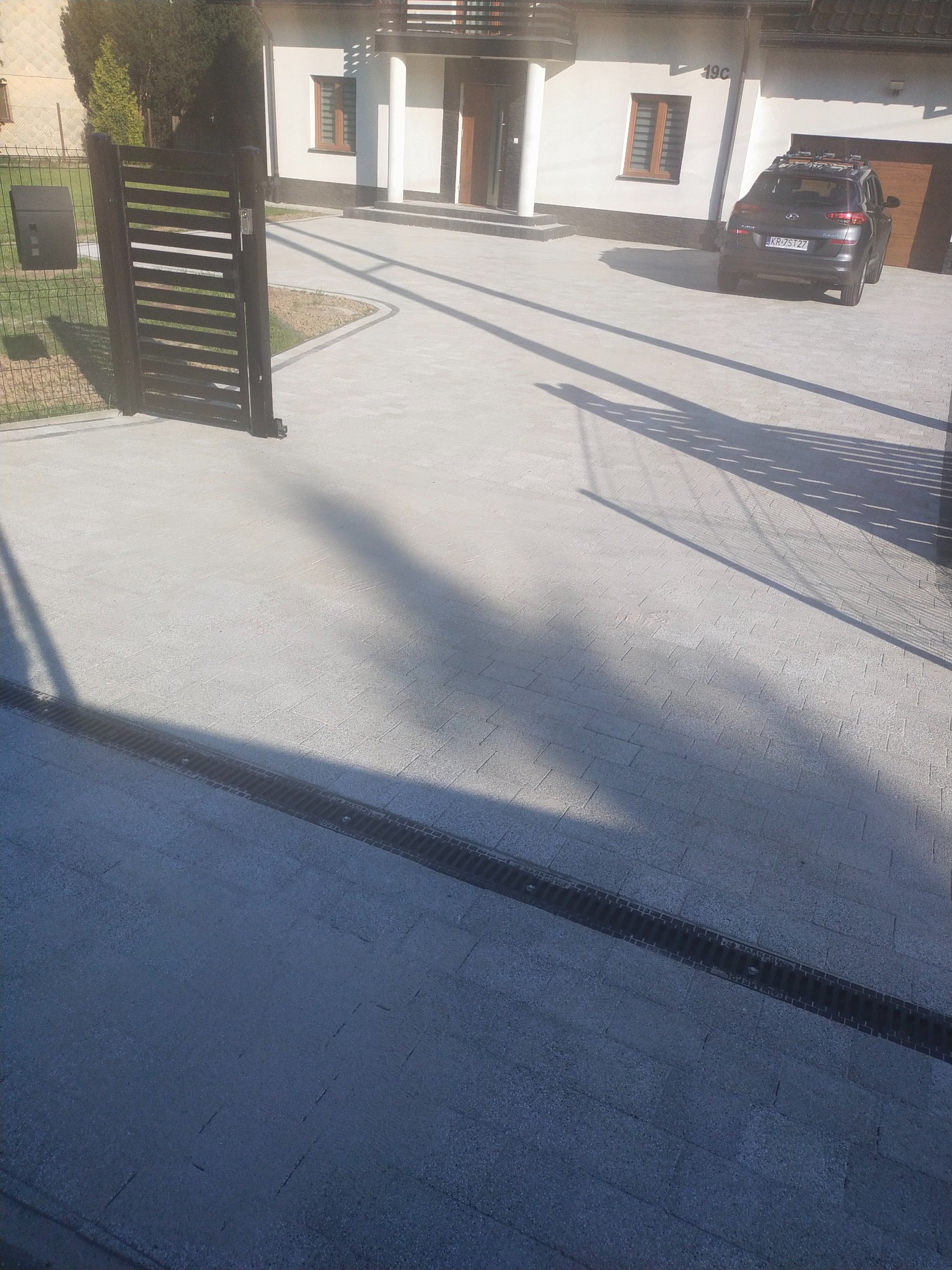 Nowoczesna posesja z szarą kostką brukową, odpływem liniowym i metalową bramą wjazdową, na tle domu z białymi kolumnami.