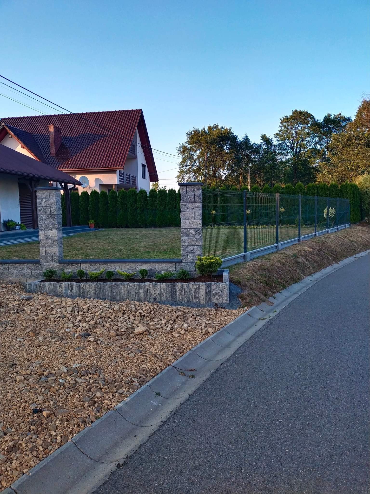 Nowoczesne ogrodzenie panelowe w kolorze grafitowym, osadzone na betonowej podmurówce, z kamiennymi słupkami, widok z boku na tle domu i zielonych tuj