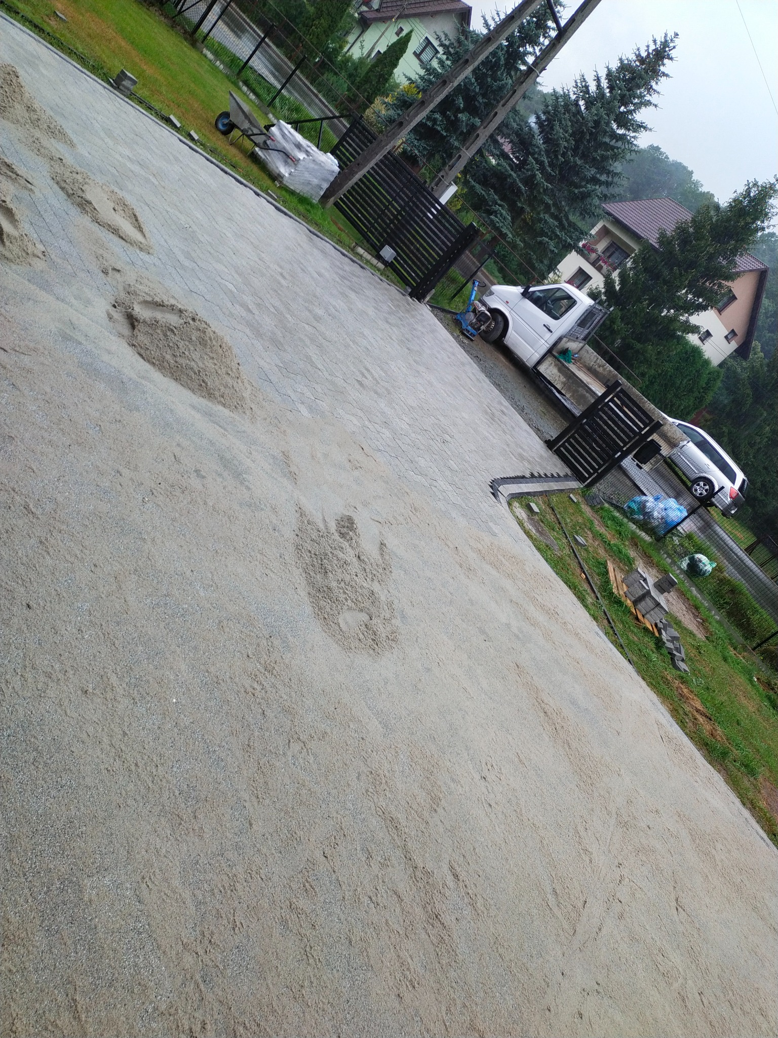 Przygotowany podkład piaskowy pod kostkę brukową, widoczne ślady po rozsypywaniu piasku, w tle brama wjazdowa i zaparkowane samochody.