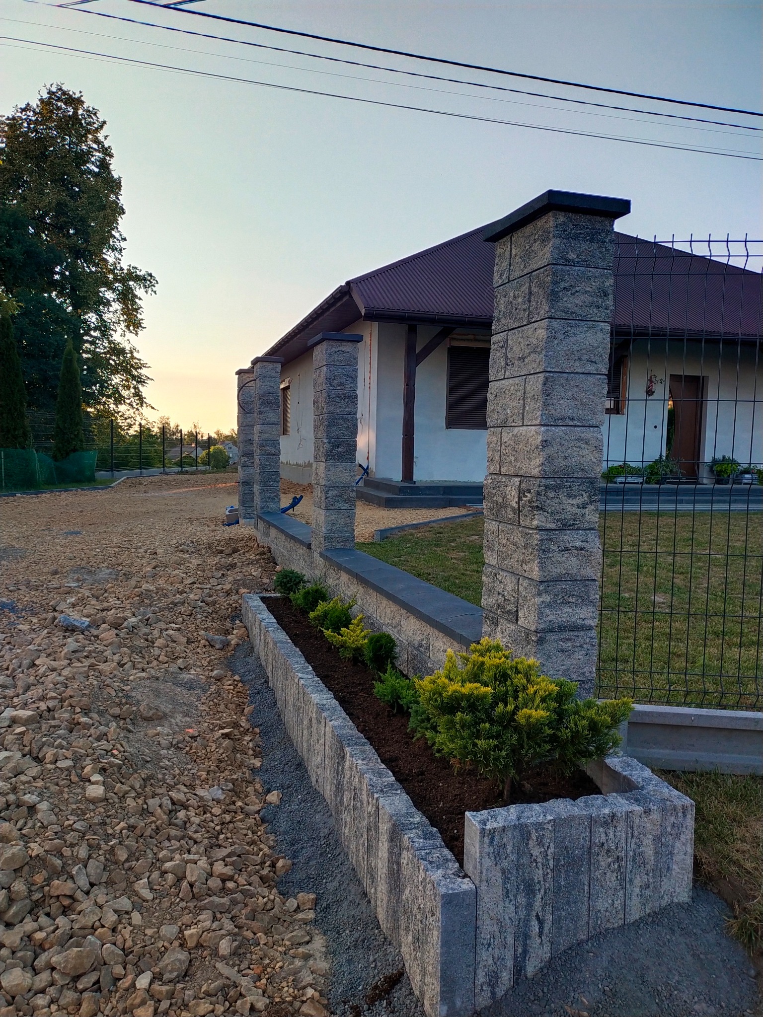 Kamienna rabata z roślinami i słupki ogrodzeniowe z szarego kamienia na tle domu jednorodzinnego w trakcie prac wykończeniowych podjazdu.