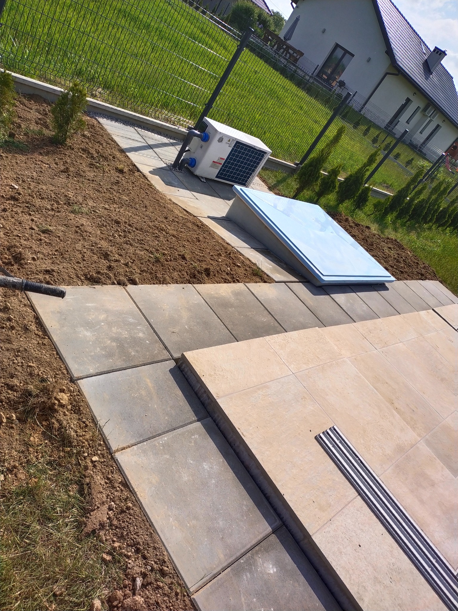 Ułożona kostka brukowa w odcieniach szarości i beżu, z widocznym odpływem liniowym, obok przygotowane podłoże pod trawnik, w tle pompa ciepła i właz basenowy, całość otoczona siatką ogrodzeniową.