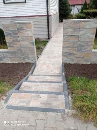 Schody i chodnik wyłożone kostką brukową prowadzące do domu z jasną elewacją, widoczne fragmenty rabat z ziemią i trawa.