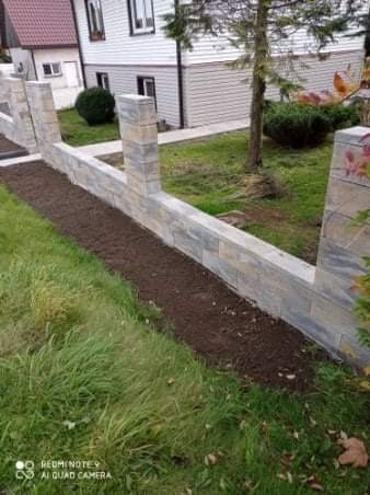 Niski murek z szarej kostki łupanej, oddzielający trawnik od rabaty z ziemią, widok na dom z białymi panelami.