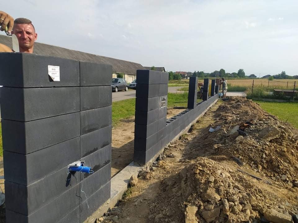Wznoszenie ogrodzenia modułowego z szarych bloczków betonowych; widoczna instalacja elektryczna w słupku oraz pracownicy podczas montażu w słoneczny dzień.