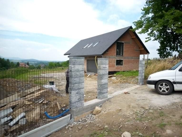 W trakcie wznoszenia ogrodzenia panelowego z betonową podmurówką i słupkami z szarej kostki łupanej, w tle dom z garażem i widok na pagórkowaty krajobraz.
