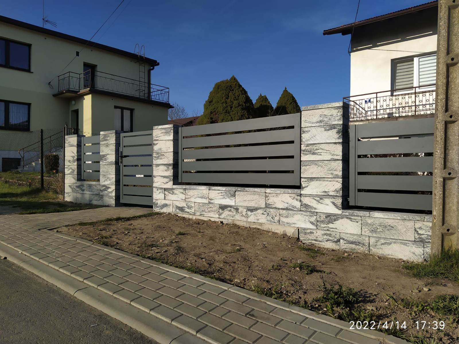 Nowoczesne, poziome, szare przęsła ogrodzeniowe zamontowane na słupkach z szarej kostki dekoracyjnej, widok z poziomu chodnika.