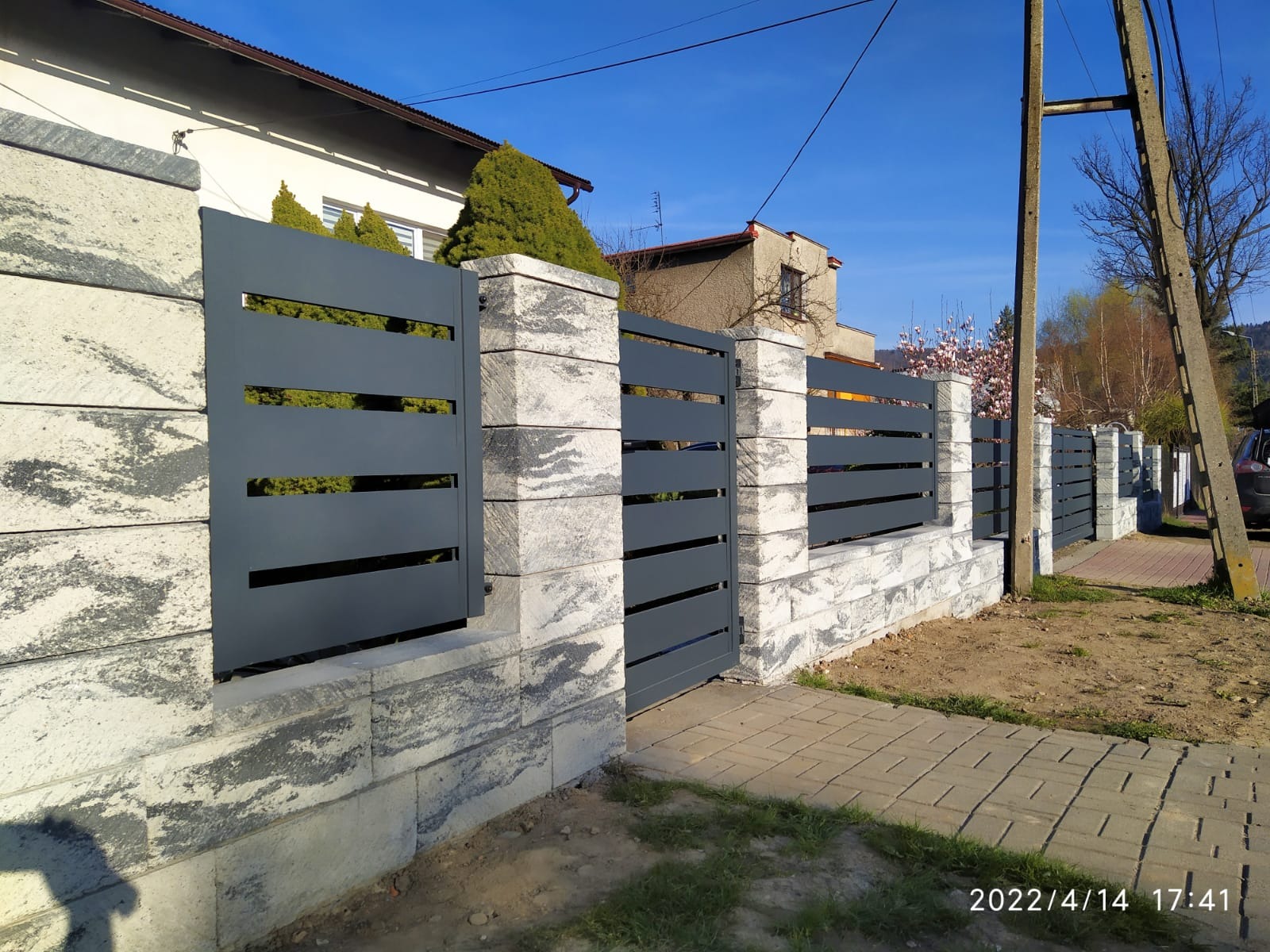 Nowoczesne ogrodzenie panelowe z poziomymi przęsłami w kolorze grafitowym, osadzone na słupkach i podmurówce z szarej kostki imitującej kamień, widoczne z poziomu chodnika w słoneczny dzień.