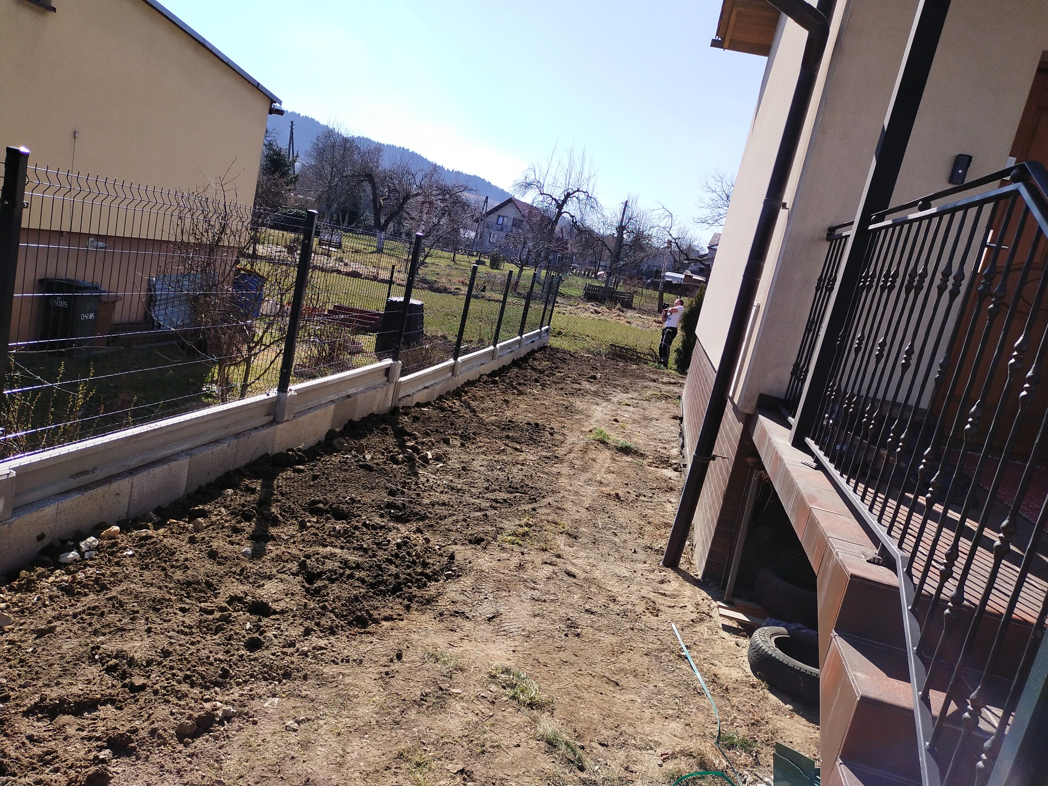 Metalowe ogrodzenie panelowe z podmurówką betonową na nierównym terenie, widok z boku domu z metalową balustradą, w tle wzgórza i zabudowania.