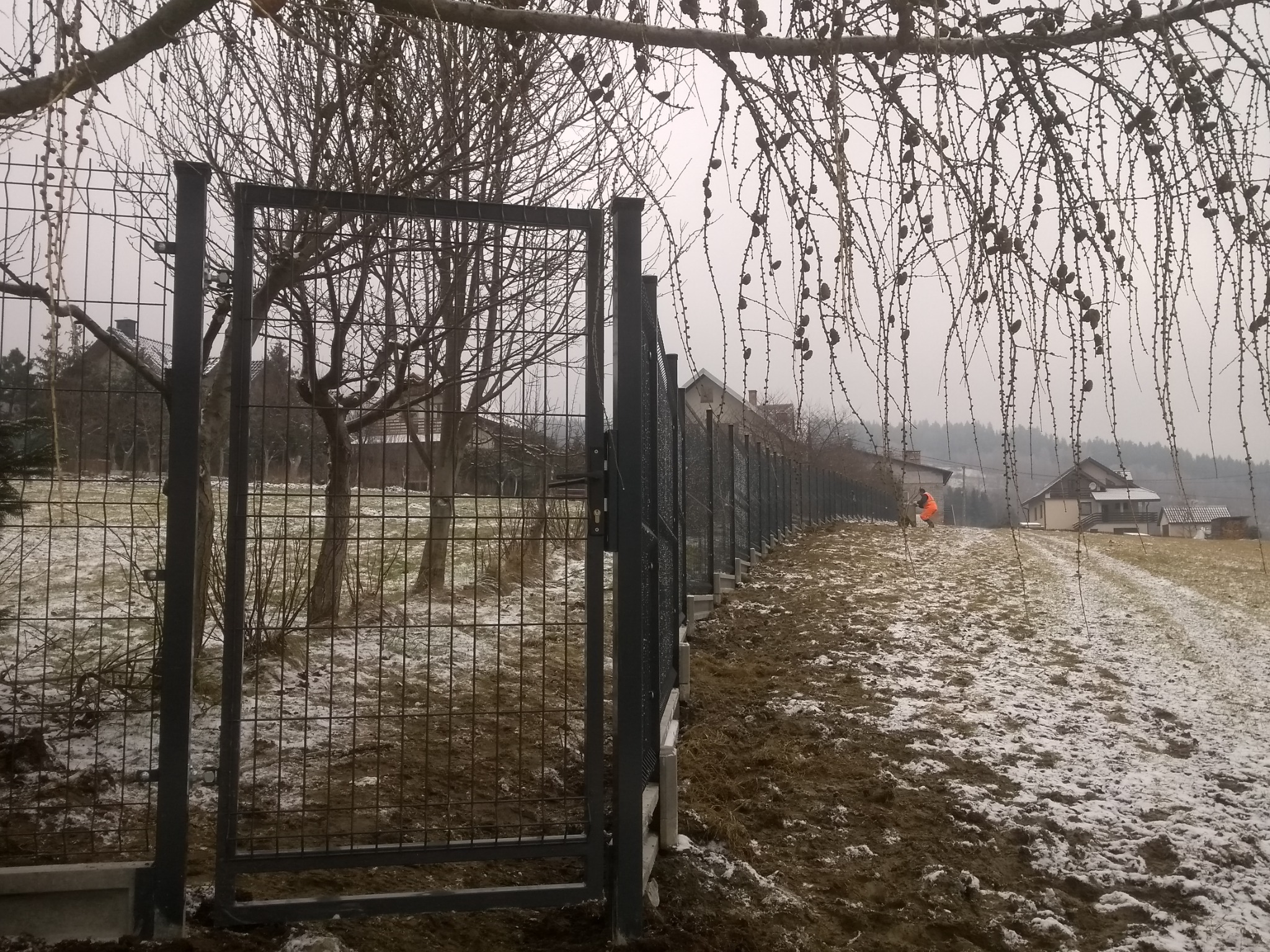 Nowo zamontowane, metalowe ogrodzenie panelowe z otwartą furtką w zimowym krajobrazie; w tle widoczny pracownik w pomarańczowym kombinezonie.