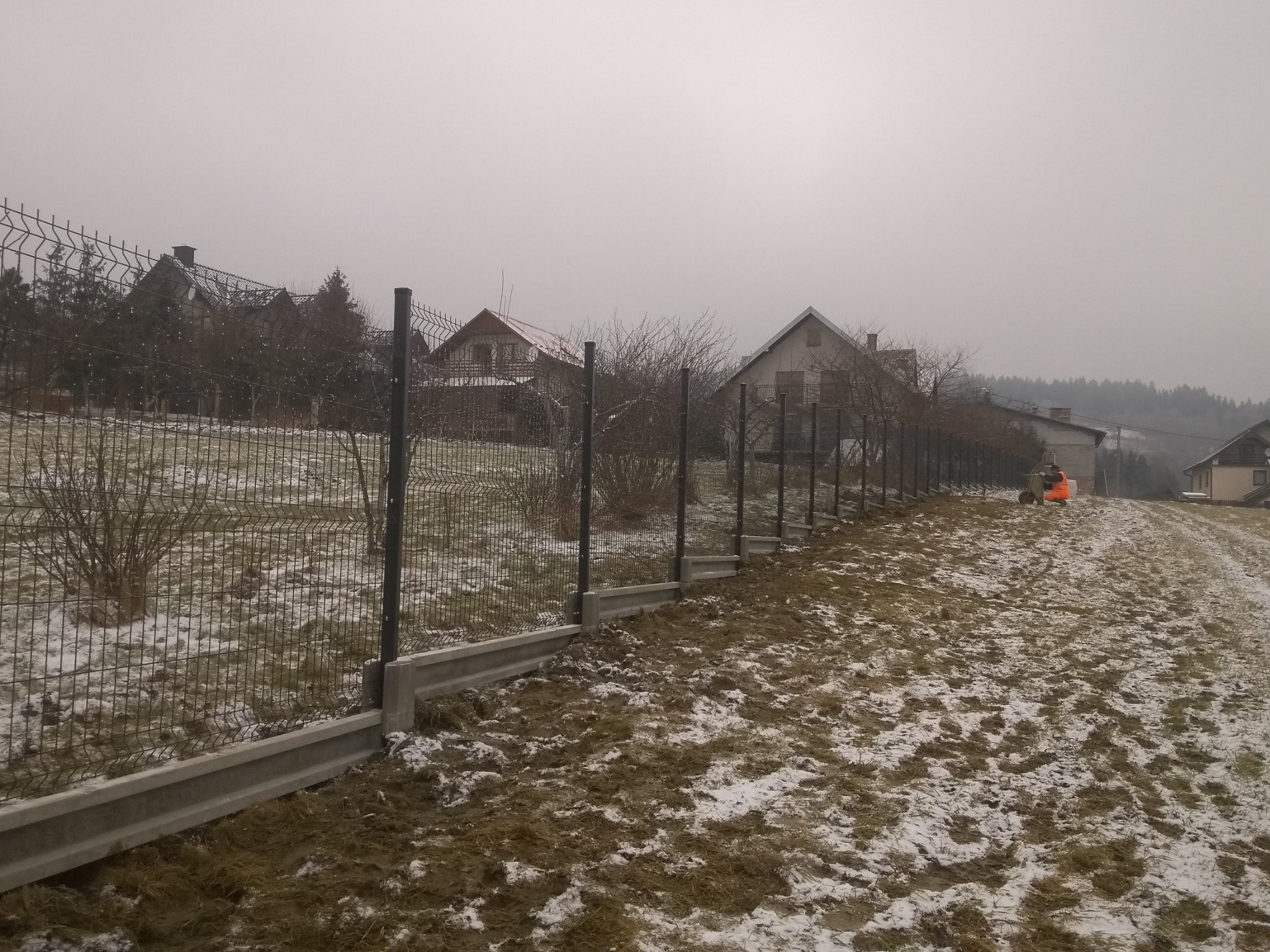 Nowoczesne ogrodzenie panelowe z podmurówką betonową, widok z zewnątrz posesji w zimowej scenerii, z fragmentami śniegu na trawie i pochmurnym niebem w tle.
