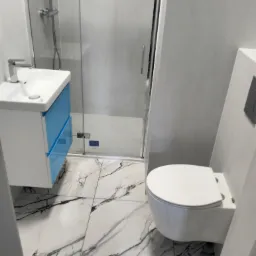 Nowoczesna łazienka z marmurowymi płytkami podłogowymi, kabiną prysznicową ze szklanymi drzwiami, umywalką z niebieskimi szufladami i podwieszaną toaletą.