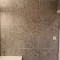 Ściana wyłożona szarymi płytkami imitującymi kamień z widoczną białą fugą, fragmentem białej szafki i podwójnym włącznikiem światła.