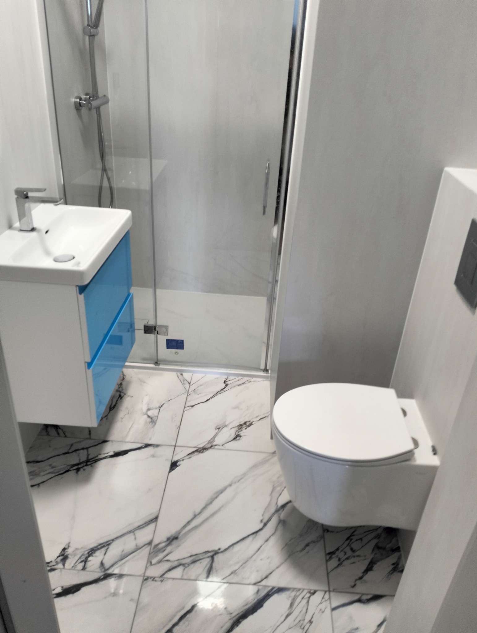 Nowoczesna łazienka z marmurowymi płytkami podłogowymi, kabiną prysznicową ze szklanymi drzwiami, umywalką z niebieskimi szufladami i podwieszaną toaletą.