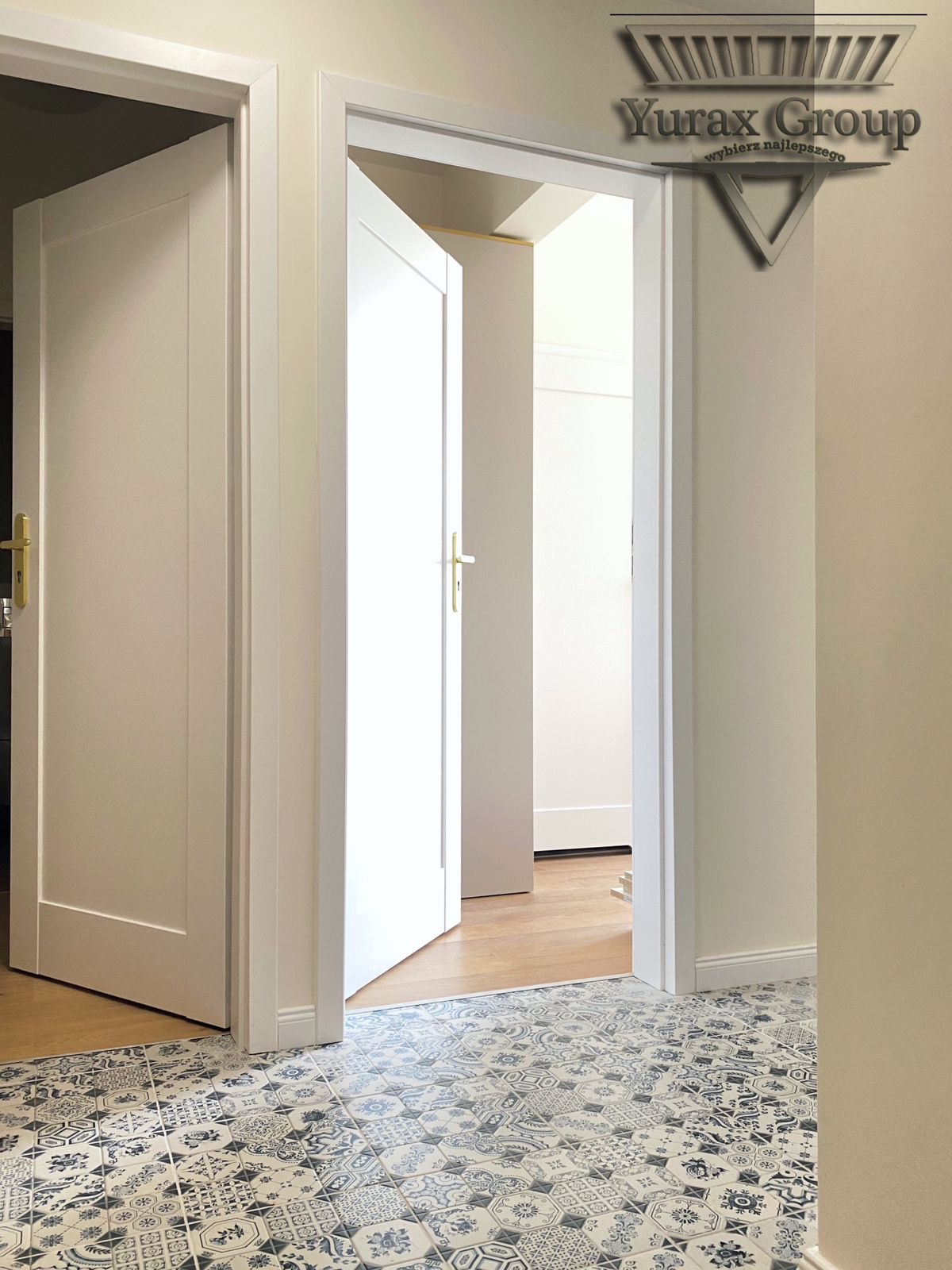 Wykończony korytarz z białymi drzwiami z złotymi klamkami i wzorzystą, niebiesko-białą podłogą z płytek, widoczne logo firmy na ścianie.