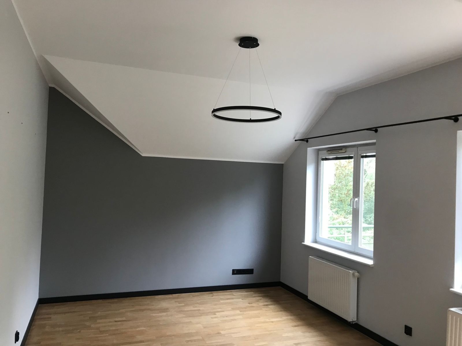 Pokój po remoncie: szara ściana, biały sufit z nietypowym załamaniem, drewniana podłoga, okno z grzejnikiem i nowoczesna okrągła lampa wisząca.