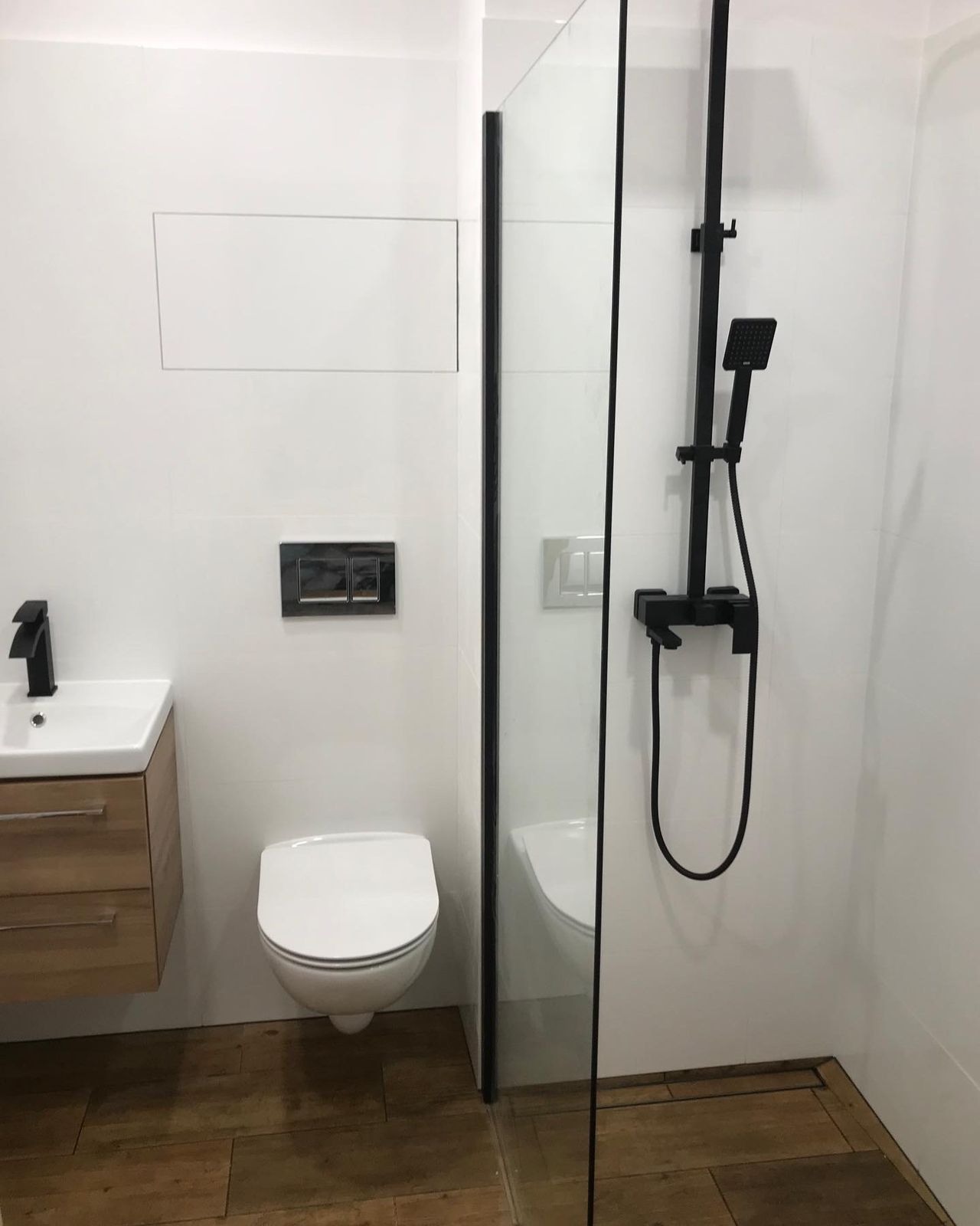 Nowoczesna łazienka z białymi płytkami, drewnopodobną podłogą, wiszącą toaletą, czarną armaturą i kabiną prysznicową ze szklaną ścianą.