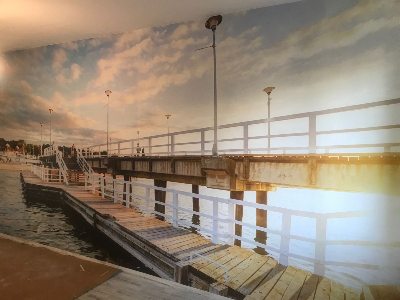 Fototapeta na ścianie z motywem molo nad morzem o zachodzie słońca, widoczne drewniane pomosty, białe balustrady i latarnie.