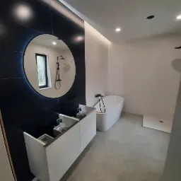 Nowoczesna łazienka z czarnymi płytkami, okrągłym lustrem z podświetleniem, białą wolnostojącą wanną i szarą podłogą z płytek w trakcie montażu szafki umywalkowej.