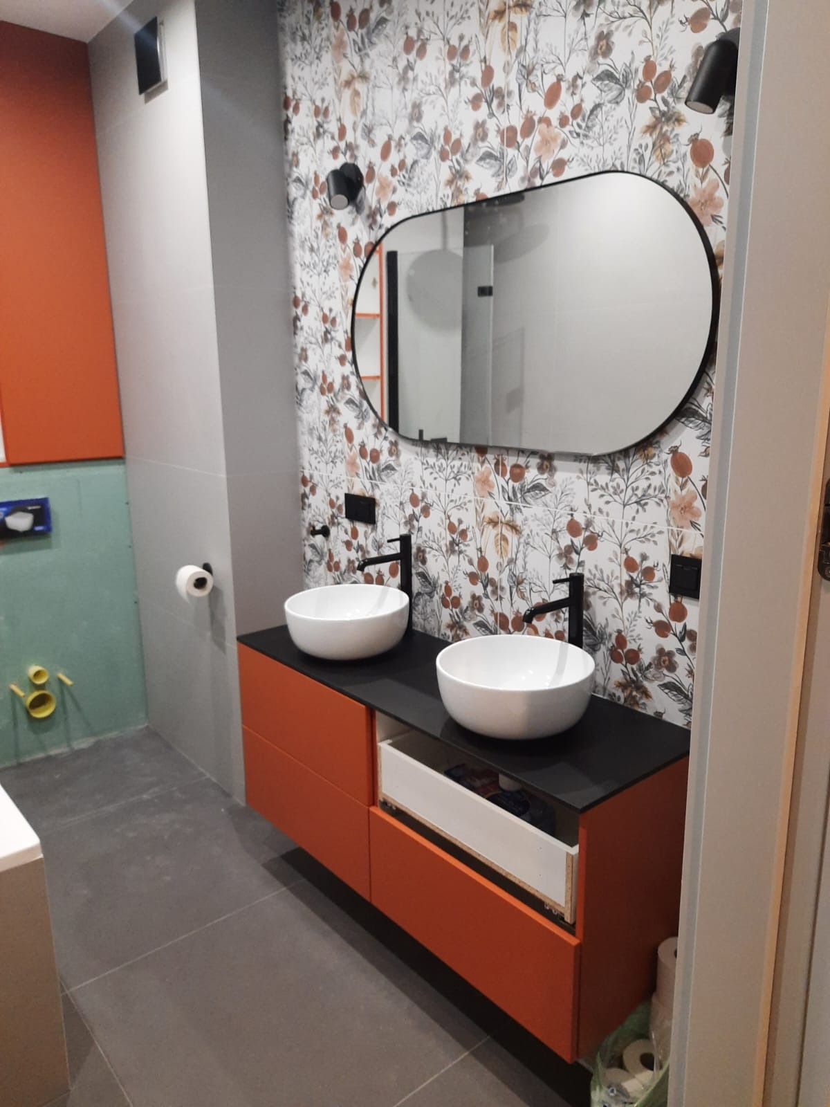 Nowoczesna łazienka z dwoma białymi umywalkami na pomarańczowej szafce z czarnym blatem, owalne lustro z czarną ramą i tapeta w kwiaty.