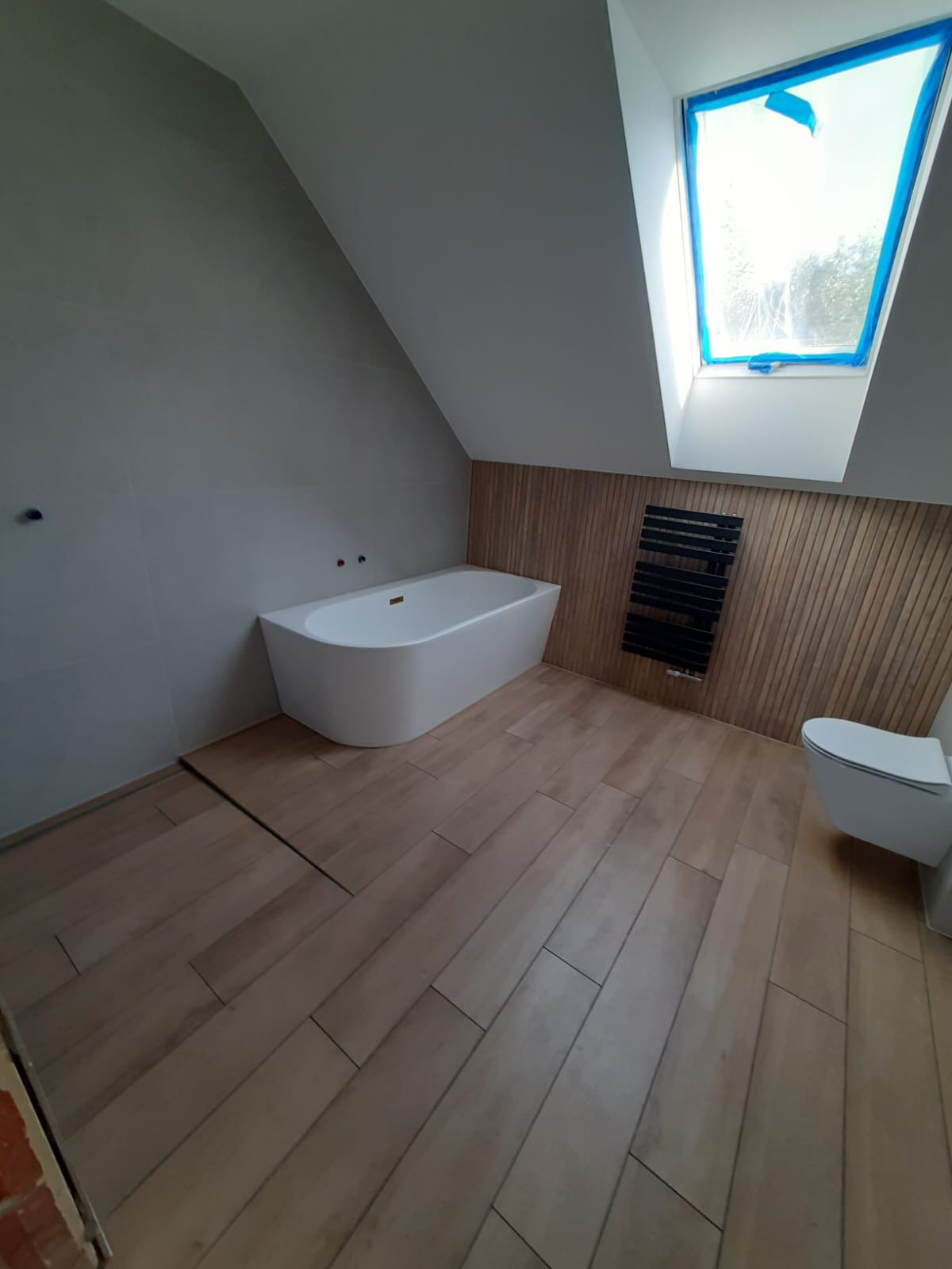 Nowoczesna łazienka na poddaszu z wolnostojącą wanną, drewnianą boazerią i płytkami imitującymi drewno na podłodze, okno dachowe zabezpieczone folią ochronną.