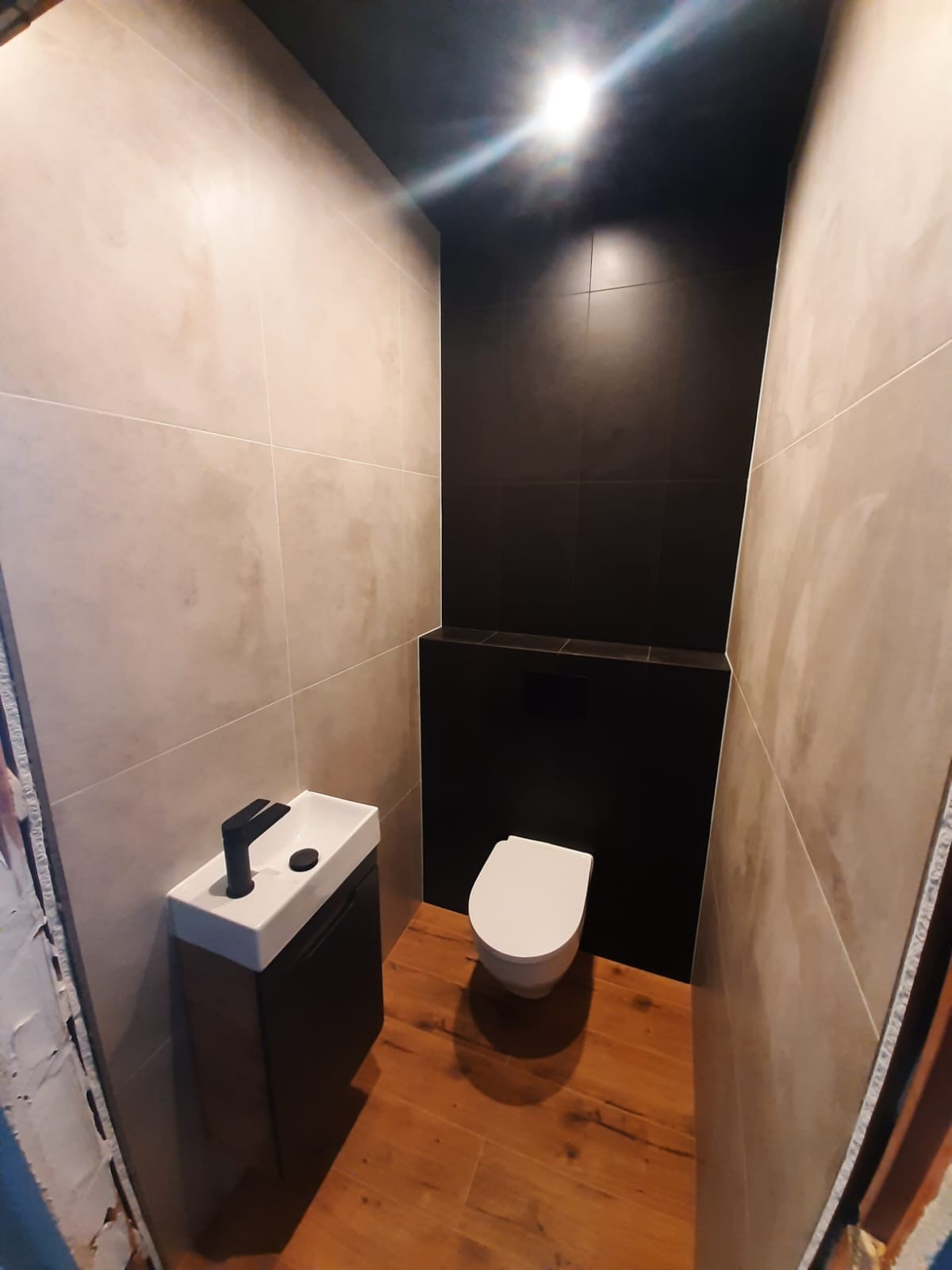 Wykończona łazienka w odcieniach beżu i czerni, z drewnianą podłogą, mała umywalka z czarną armaturą i podwieszana toaleta.