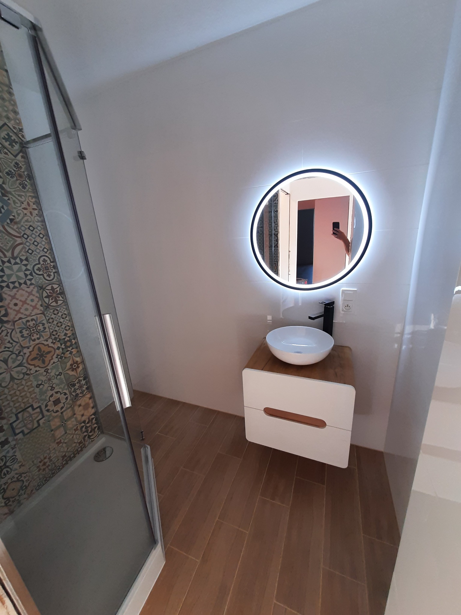 Nowoczesna łazienka z kabiną prysznicową z wzorzystymi płytkami, okrągłym lustrem z podświetleniem LED nad białą szafką umywalkową z drewnianym blatem i ceramiczną umywalką.