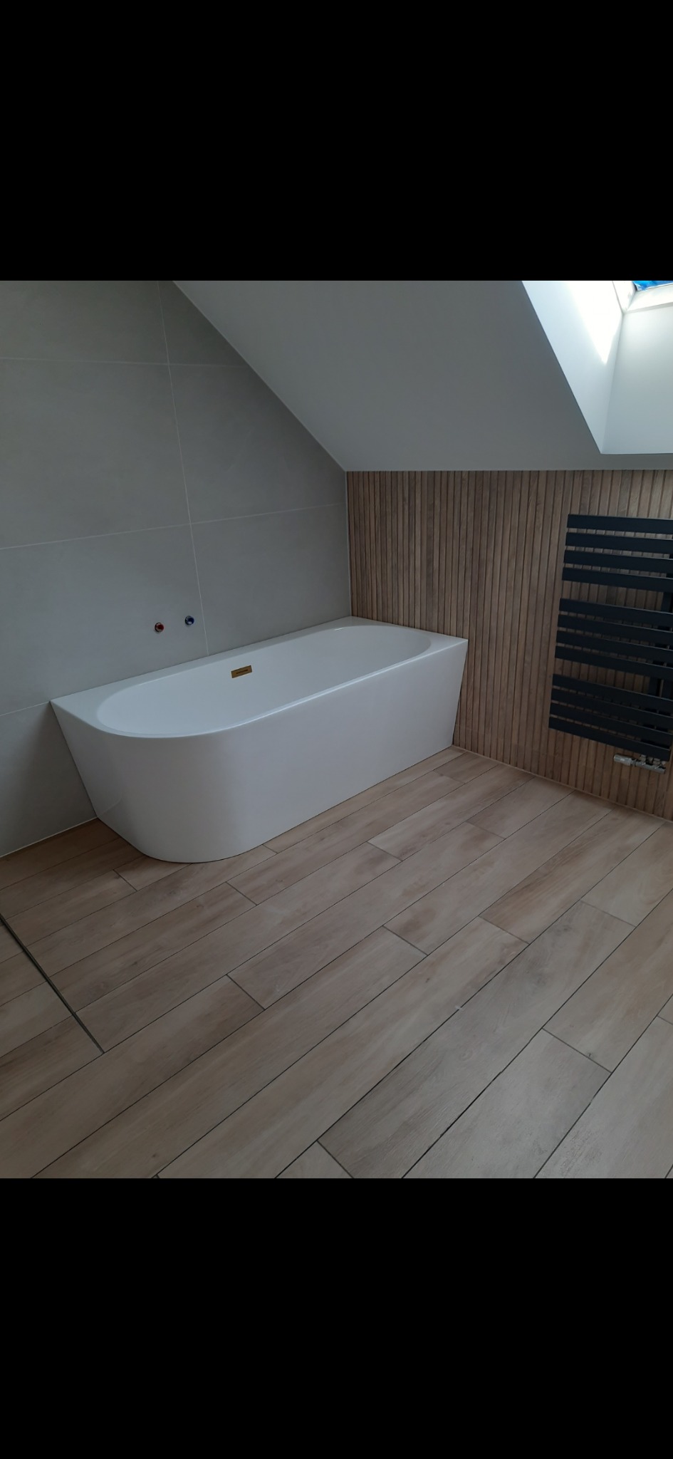 Nowoczesna łazienka na poddaszu z wolnostojącą białą wanną, grzejnikiem drabinkowym i drewnianą okładziną ściany, oświetlona światłem dziennym z okna dachowego.