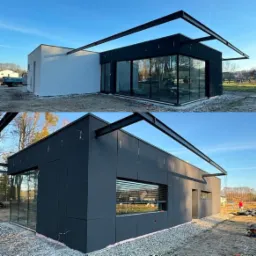 Nowoczesny dom z szarą elewacją panelową i przeszklonym narożnikiem, widoczne stalowe elementy konstrukcyjne zadaszenia, teren wokół domu w trakcie prac wykończeniowych.
