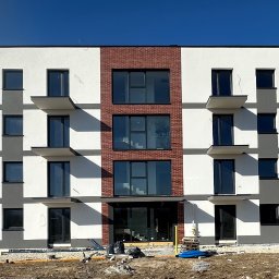 ELEBUD ELEWACJE DOCIEPLENIA - Nowoczesny budynek wielorodzinny z elewacją w odcieniach bieli, szarości i cegły, z balkonami i dużymi oknami, w słoneczny dzień. Widoczne ogrodzenie placu budowy.