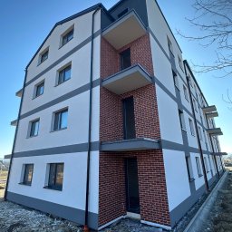 ELEBUD ELEWACJE DOCIEPLENIA - Nowoczesny budynek wielorodzinny z elewacją w odcieniach bieli i szarości, z elementami dekoracyjnymi z cegły. Widok z dołu, błękitne niebo w tle.