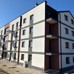 ELEBUD ELEWACJE DOCIEPLENIA - Nowoczesny budynek wielorodzinny z elewacją w odcieniach bieli, szarości i cegły, z balkonami i dużymi oknami, na tle błękitnego nieba. Widok z dołu ukazuje detale wykończenia.