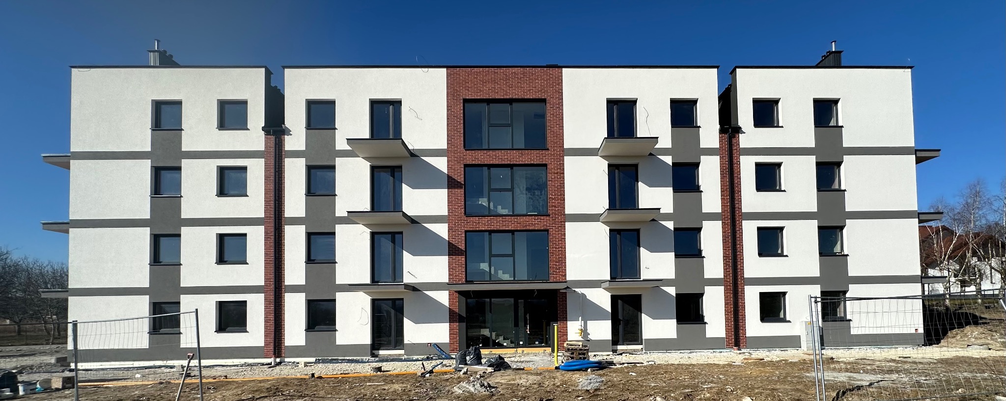 Nowoczesny budynek wielorodzinny z elewacją w odcieniach bieli, szarości i cegły, z balkonami i dużymi oknami, w słoneczny dzień. Widoczne ogrodzenie placu budowy.