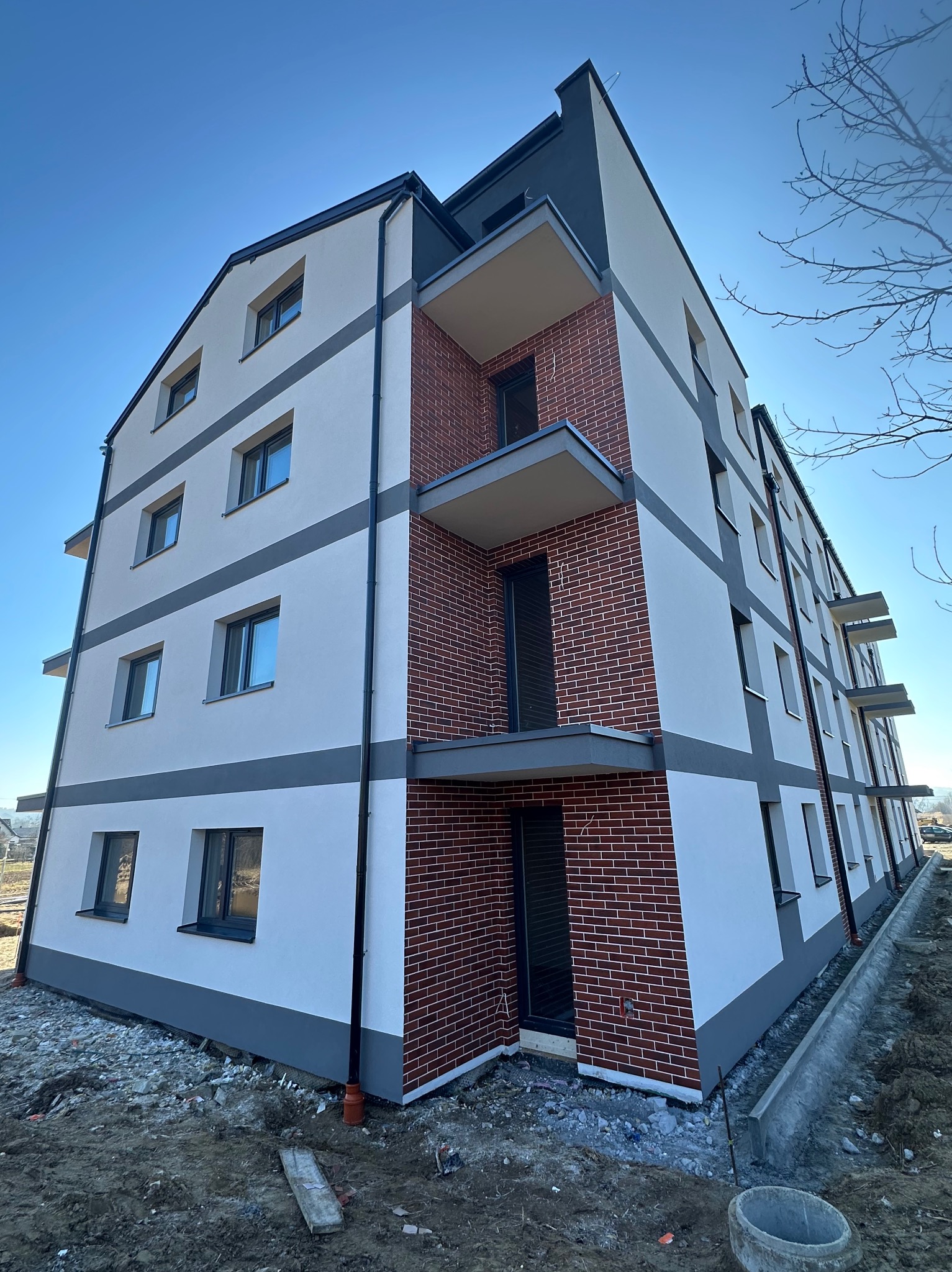 Nowoczesny budynek wielorodzinny z elewacją w odcieniach bieli i szarości, z elementami dekoracyjnymi z cegły. Widok z dołu, błękitne niebo w tle.