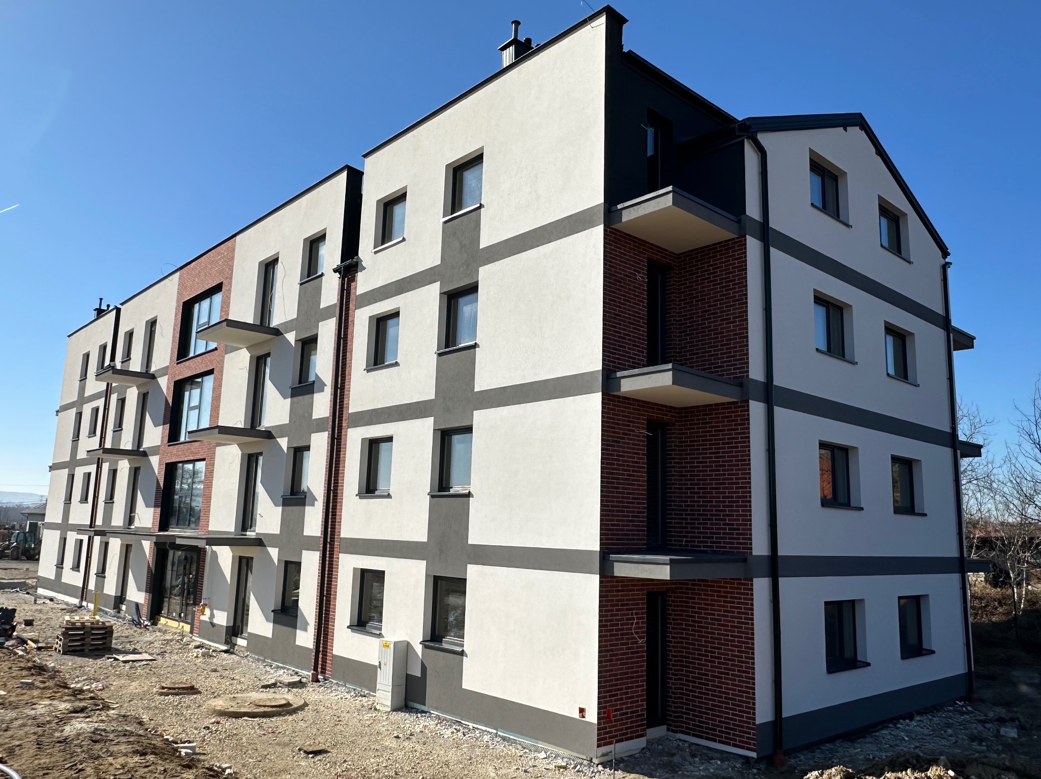 Nowoczesny budynek wielorodzinny z elewacją w odcieniach bieli, szarości i cegły, z balkonami i dużymi oknami, na tle błękitnego nieba. Widok z dołu ukazuje detale wykończenia.