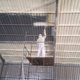 Pimar Izolacje S.C - Pracownik w kombinezonie ochronnym aplikuje pianę ZK na metalowy dach z rusztowania, widoczne fragmenty dachu już pokryte pianą.