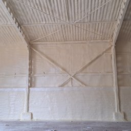 Pimar Izolacje S.C - Wnętrze hali przemysłowej po natrysku izolacji pianką na ściany i dach, widoczne stalowe elementy konstrukcyjne pokryte pianą, nierówna powierzchnia izolacji.