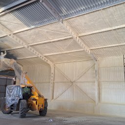 Pimar Izolacje S.C - Aplikacja izolacji pianką na metalowej konstrukcji hali, pracownik w kombinezonie na podnośniku, widoczne ślady piany na elementach konstrukcyjnych i ścianach.