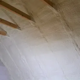 Poddasze z drewnianą konstrukcją pokryte warstwą jasnej, natryskiwanej piany izolacyjnej, widoczne bloczki betonowe i okno.