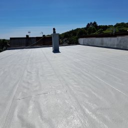 Pimar Izolacje S.C - Jasnoszary, płaski dach po renowacji z widocznym kominem i otaczającą zabudową w tle, wykonany w słoneczny dzień.