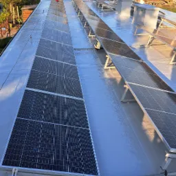 Widok z góry na dach pokryty jasnoszarą, prawdopodobnie żywiczną powłoką, na którym zamontowane są panele słoneczne w rzędach, z widocznymi elementami montażowymi i otaczającą zielenią drzew...