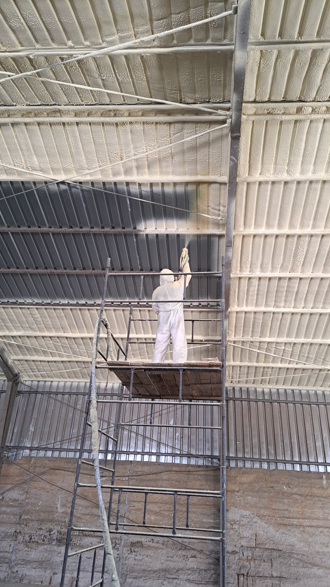 Pracownik w kombinezonie ochronnym aplikuje pianę ZK na metalowy dach z rusztowania, widoczne fragmenty dachu już pokryte pianą.