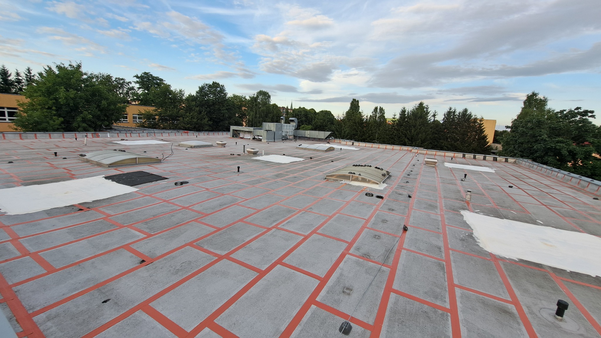 Płaski dach budynku pokryty szarą papą z czerwonymi liniami podziału, widoczne świetliki, kominy wentylacyjne i fragmenty białej powłoki ochronnej.