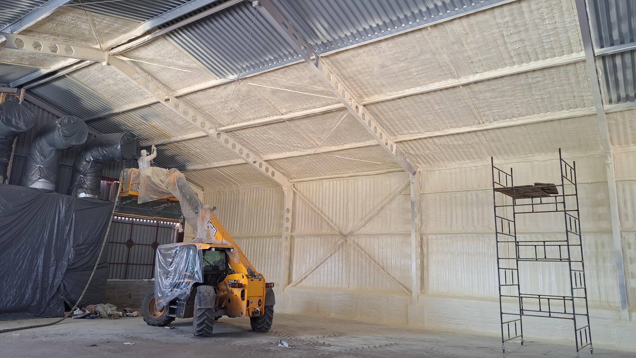 Aplikacja izolacji pianką na metalowej konstrukcji hali, pracownik w kombinezonie na podnośniku, widoczne ślady piany na elementach konstrukcyjnych i ścianach.