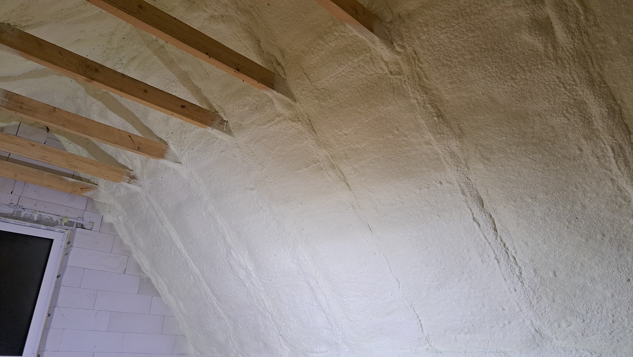 Poddasze z drewnianą konstrukcją pokryte warstwą jasnej, natryskiwanej piany izolacyjnej, widoczne bloczki betonowe i okno.