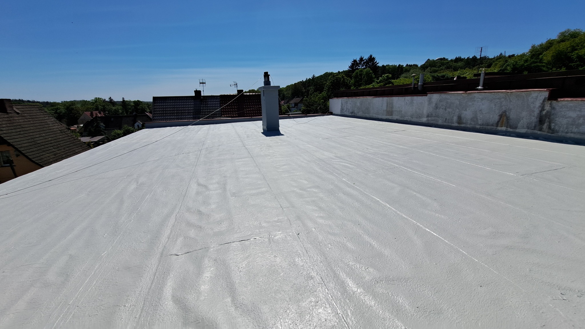 Jasnoszary, płaski dach po renowacji z widocznym kominem i otaczającą zabudową w tle, wykonany w słoneczny dzień.