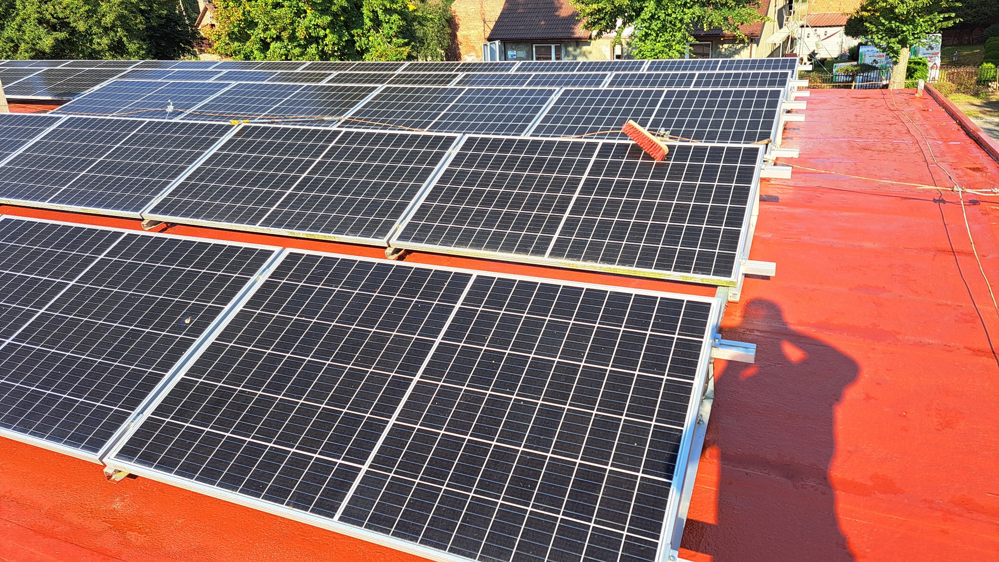 Montaż paneli fotowoltaicznych na czerwonym dachu, widoczny cień osoby oraz szczotka do czyszczenia paneli.