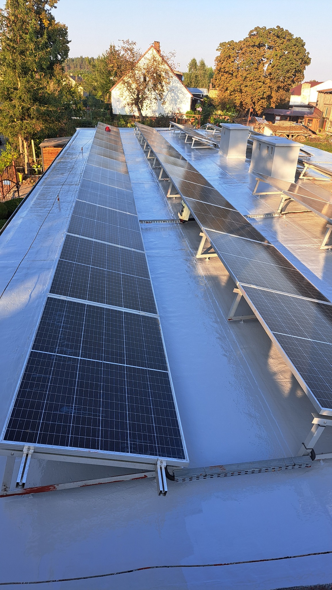 Widok z góry na dach pokryty jasnoszarą, prawdopodobnie żywiczną powłoką, na którym zamontowane są panele słoneczne w rzędach, z widocznymi elementami montażowymi i otaczającą zielenią drzew...