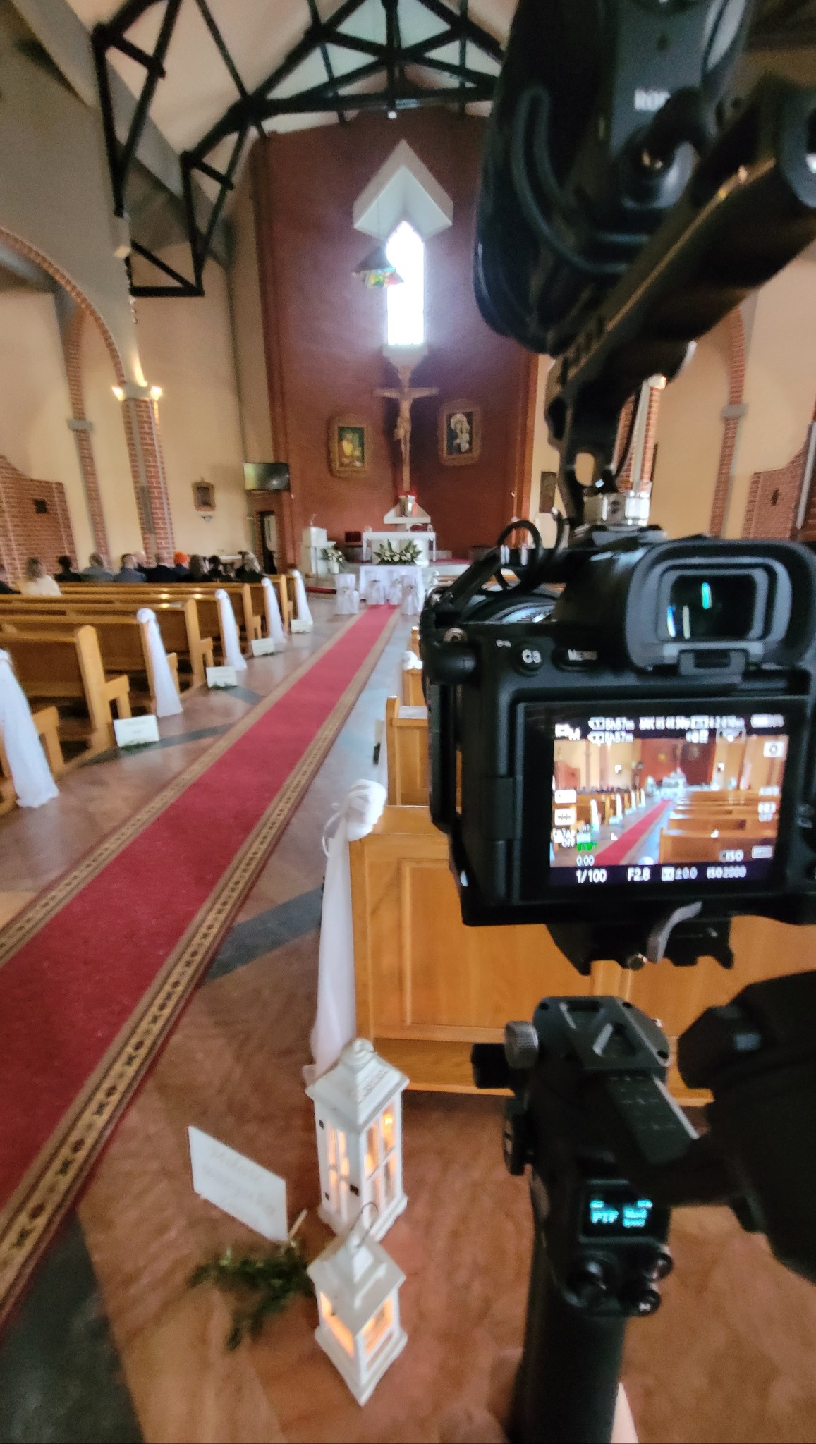Kamera z mikrofonem i stabilizatorem nagrywa wnętrze kościoła przygotowanego do ceremonii ślubnej, widoczny czerwony dywan, dekoracje z białych lampionów i kwiatów.